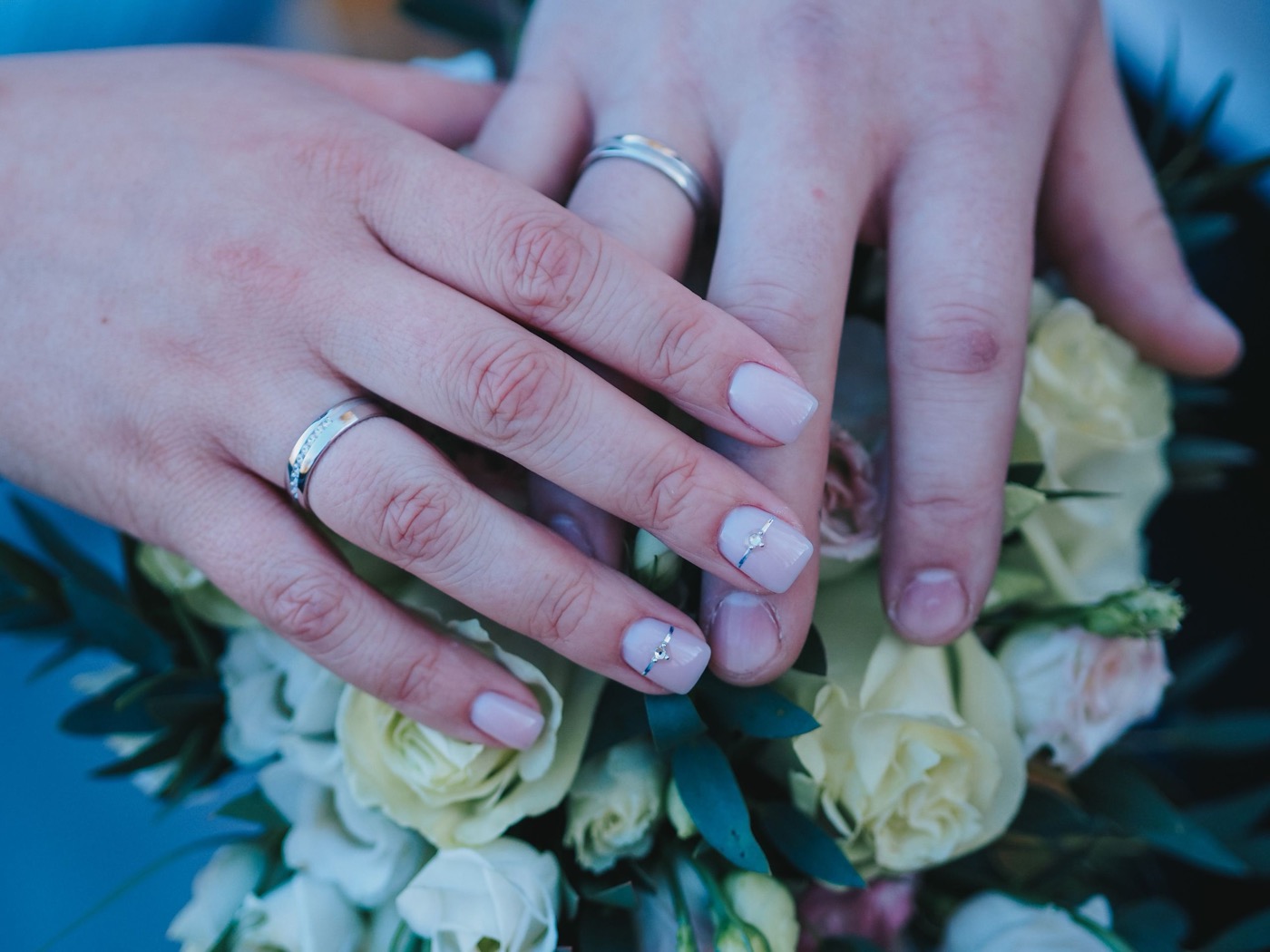 A married couple's hands resting together over a white rose bouquet, both wearing wedding bands.