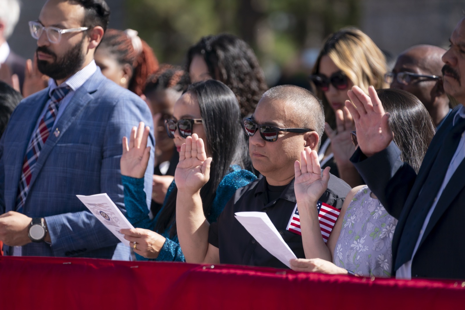 Naturalization applicants standing with their right hands raised, holding small United States flags, as they take the Oath of Allegiance at an outdoor ceremony.